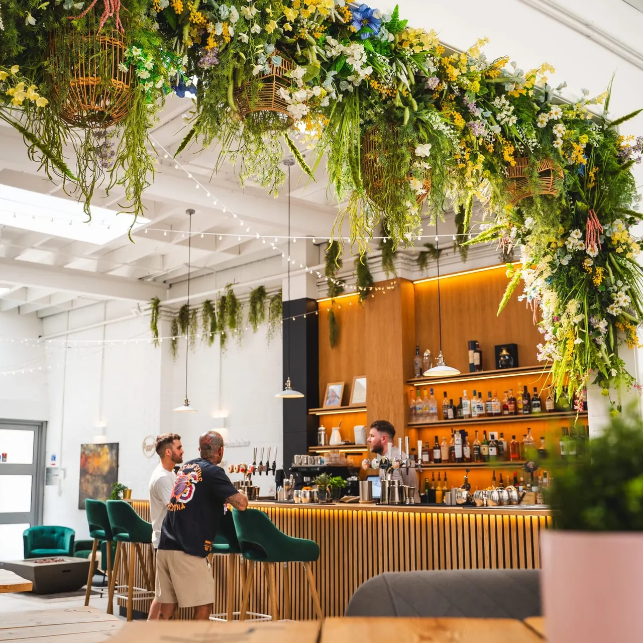 Modern bar interior with hanging baskets of yellow, white and blue flowers and trailing greenery, staff and customers at an orange-fronted counter with bottle displays.