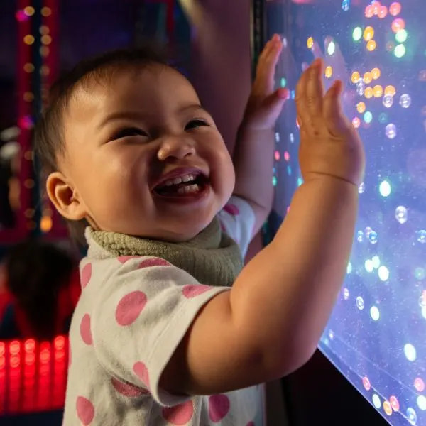 A smiling toddler reaches towards an interactive light display with colourful glowing bubbles.