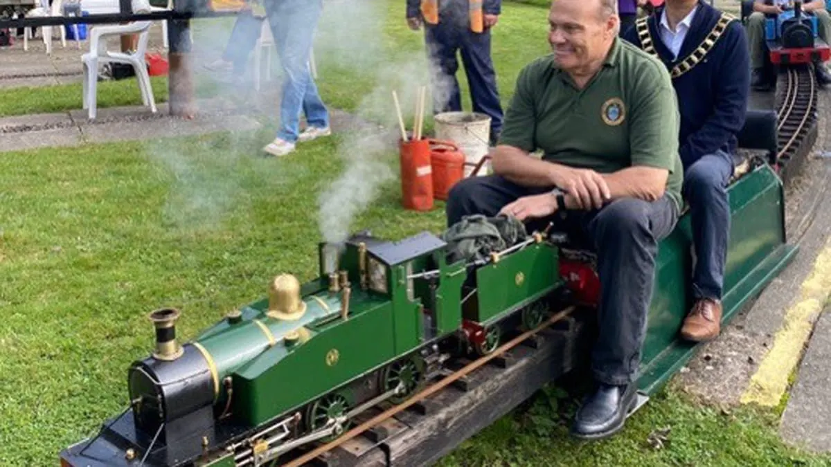 Man operating a miniature steam locomotive on a garden railway track, with spectators watching nearby.