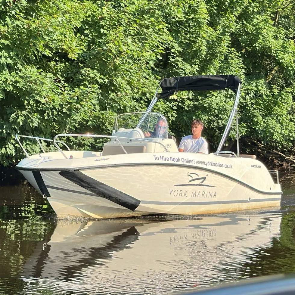 A white motorboat with a black canopy moored on calm water surrounded by overhanging green trees, with a man standing on deck.