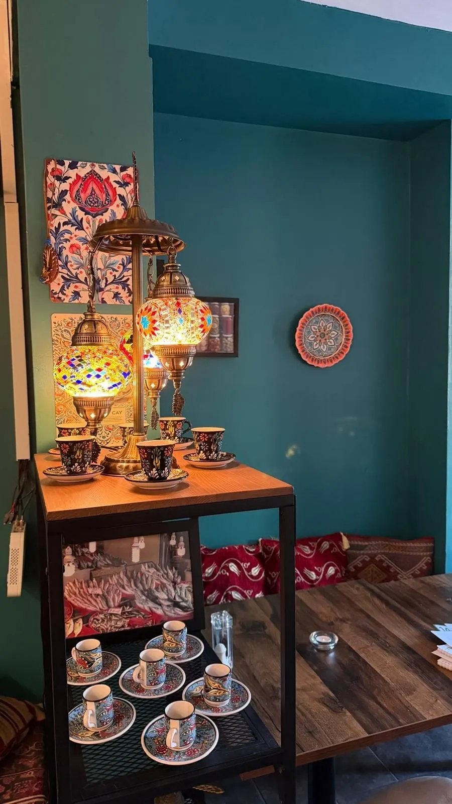 Display of Turkish decorative items including ornate lantern, patterned textiles, and traditional coffee cups on a wooden table against a teal wall.
