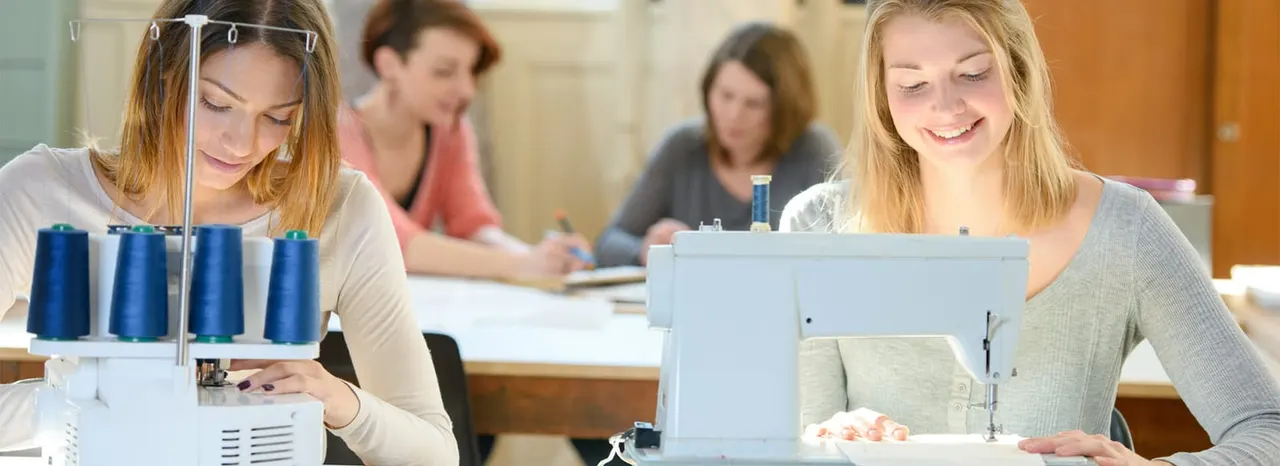 Women work at sewing machines during a class, with others seated at tables in the background.