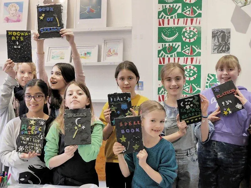 Group of children holding handmade spell books and artwork in an art studio or creative workshop space.