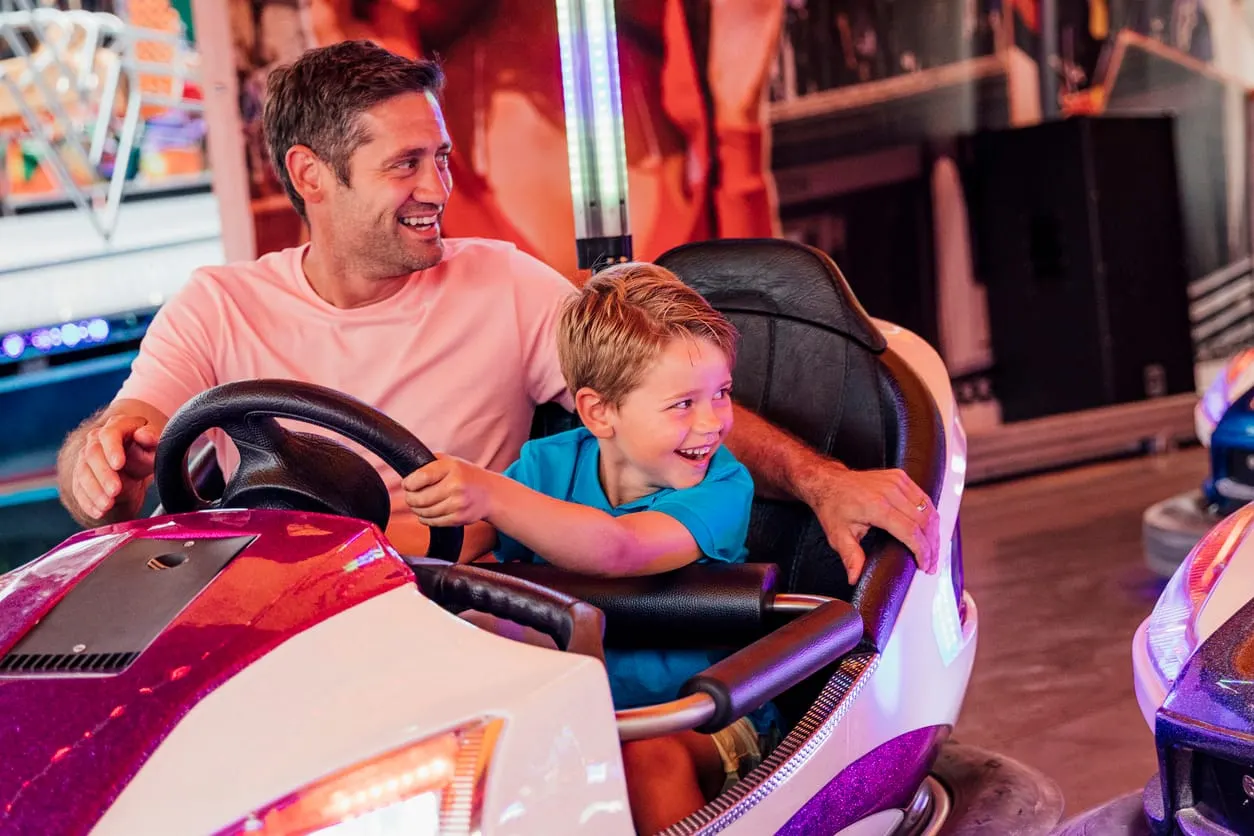 Man and young child laughing whilst driving a bumper car in an arcade setting with colourful lights.
