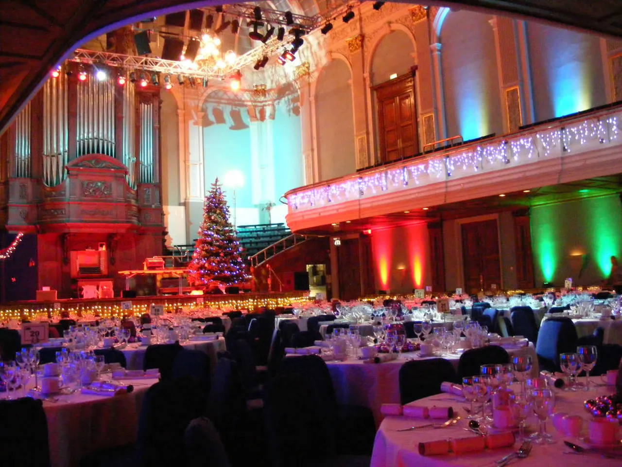 Grand concert hall decorated for a festive event with round tables, colourful lighting, a Christmas tree, and ornate architectural features including an organ and balcony.