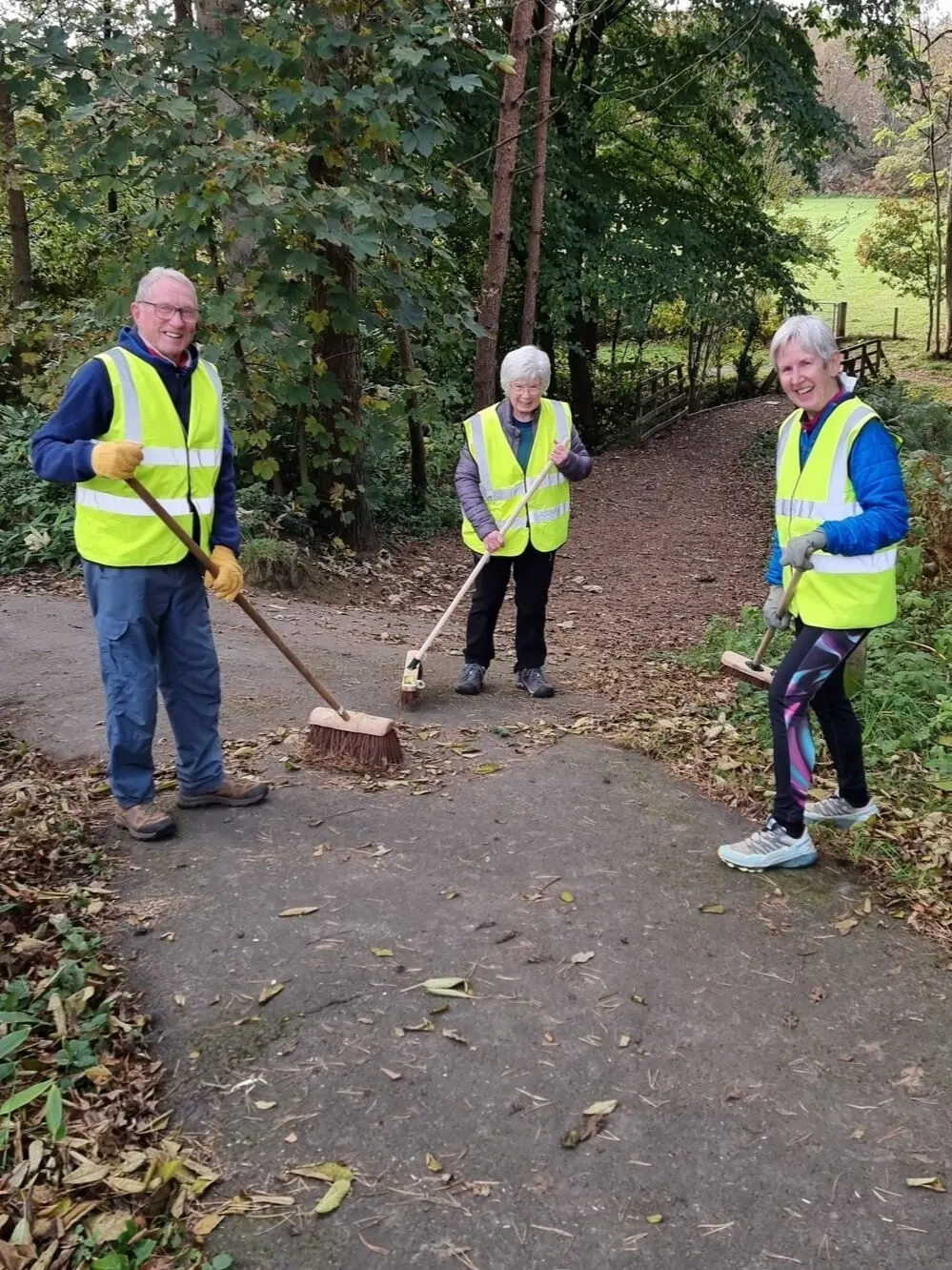 Three volunteers wearing high-visibility vests maintain a woodland path with tools at Plean Country Park.