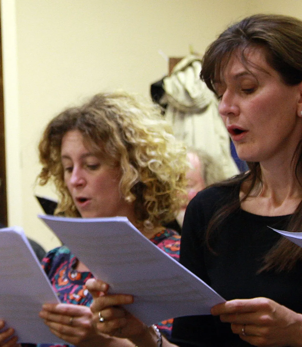 Two women singing from sheet music indoors, with a dog visible in the background.