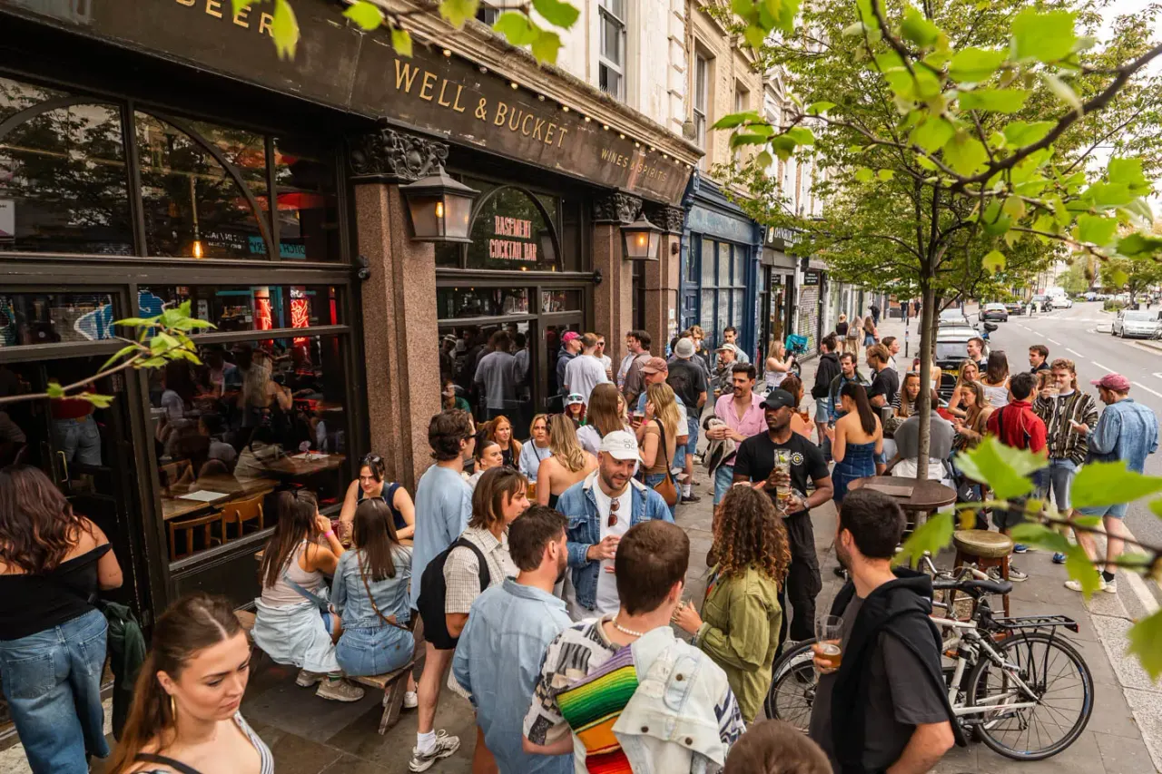Crowded street scene outside a bar with many people socialising on the pavement, some holding drinks.