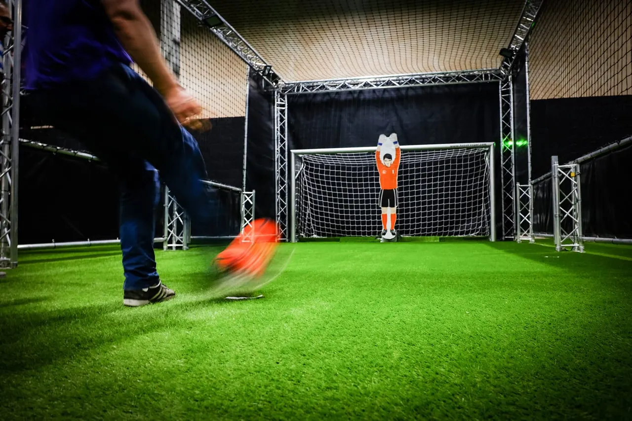 Person kicking a football towards a goalkeeper in an orange shirt standing in front of a goal net indoors.