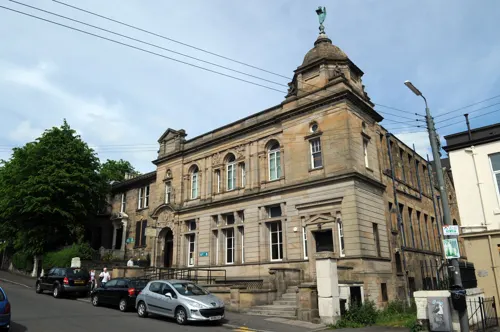 Historic sandstone library building with distinctive turret and multiple storeys, parked cars on street below.
