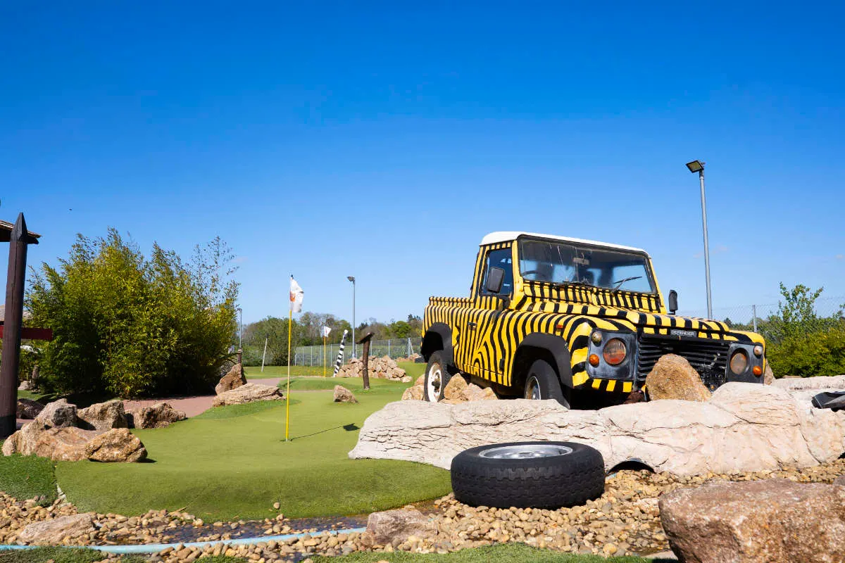 A yellow-and-black striped vehicle on a mini golf course with rocks, boulders, and a tyre obstacle.