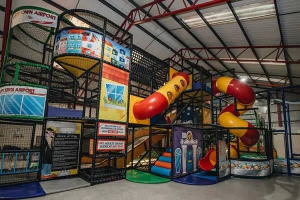 Indoor play structure with colourful tubes, slides and climbing equipment in a large warehouse-style facility.
