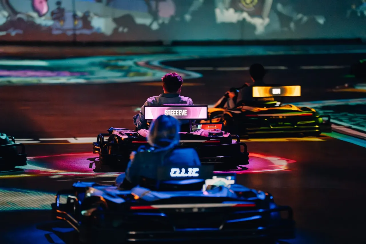 Multiple racers in electric go-karts on an indoor track with neon lighting and a large screen displaying a player name.