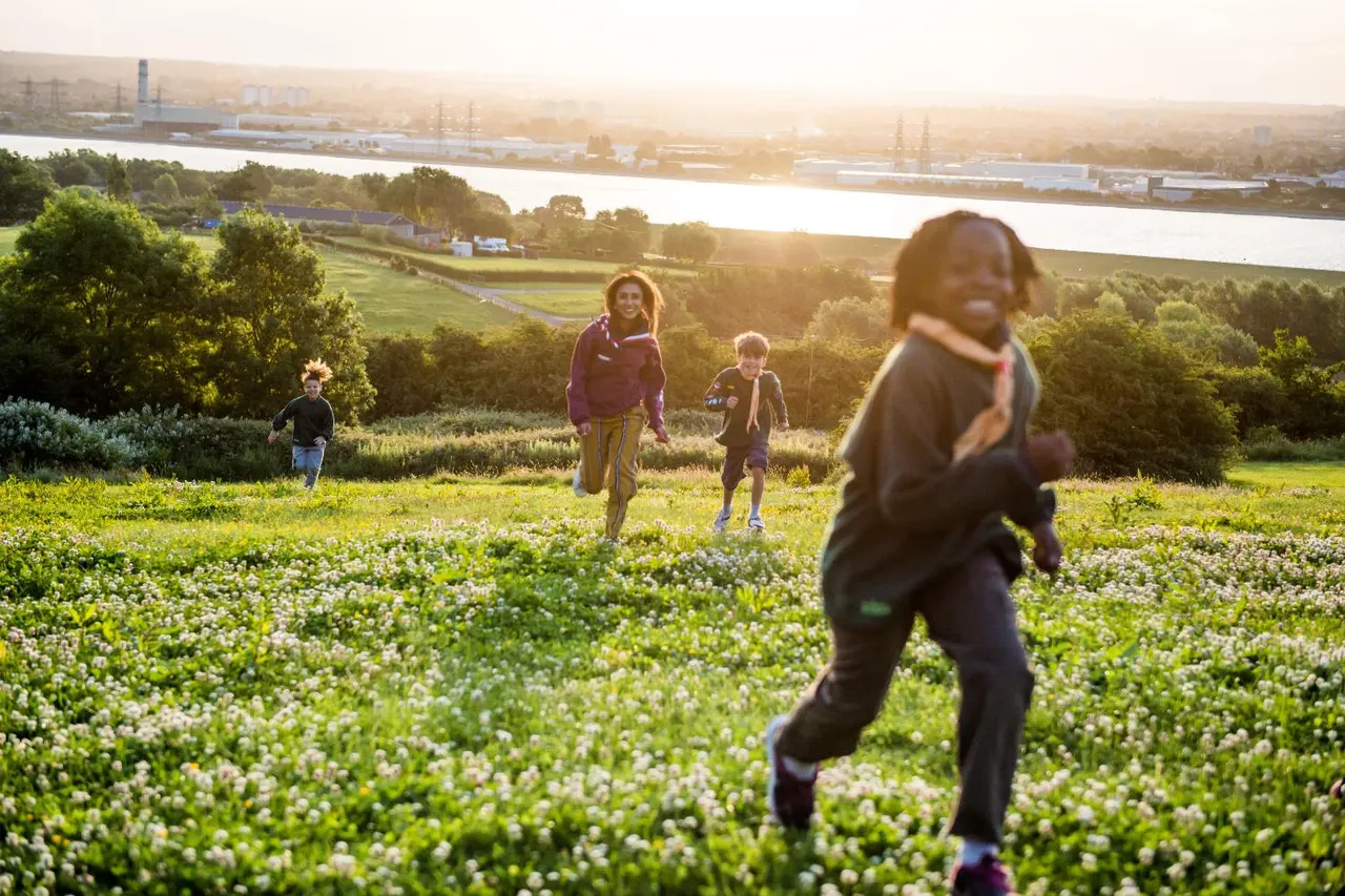 Children running through a wildflower meadow with a river and industrial landscape visible in the distance.
