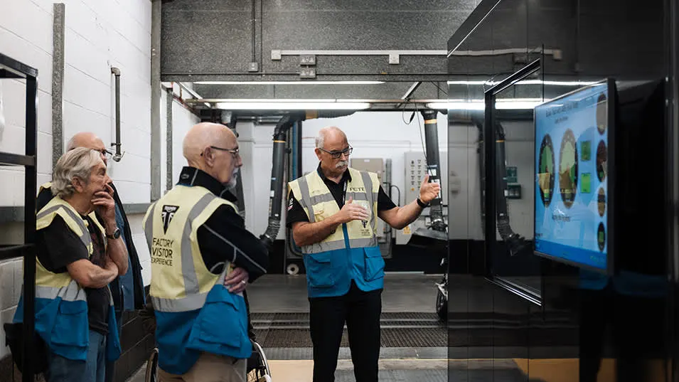 Four adults wearing hi-visibility vests watch a presentation on a screen inside an industrial factory building.