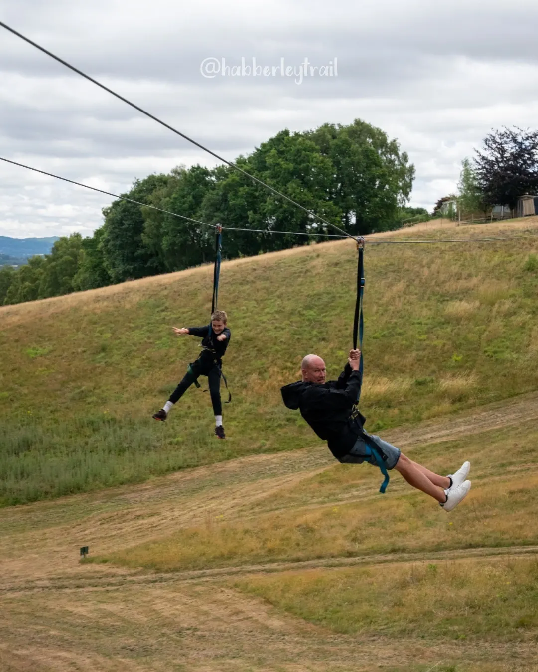 Two people zip-lining down a hillside over grassy fields with trees and mountains visible in the background.
