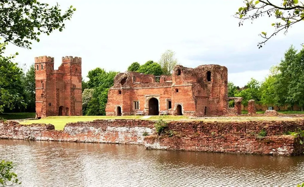 Ruined red brick castle with towers beside a moat, surrounded by green trees and lawns.