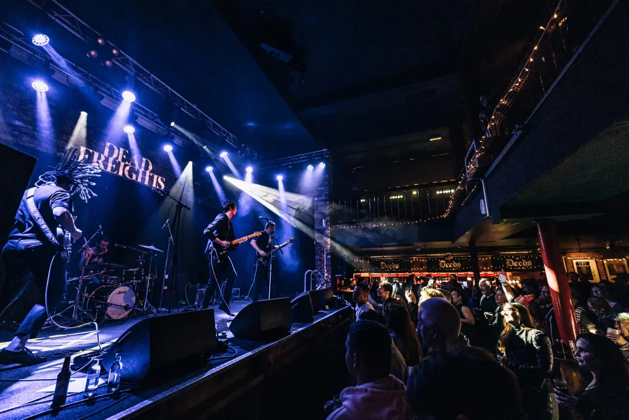 Band performing on illuminated stage with crowd watching at indoor music venue.
