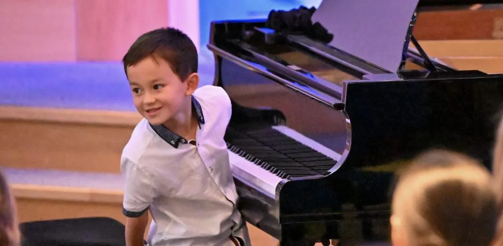 Young child in white shirt smiling next to a grand piano on stage with coloured lighting.