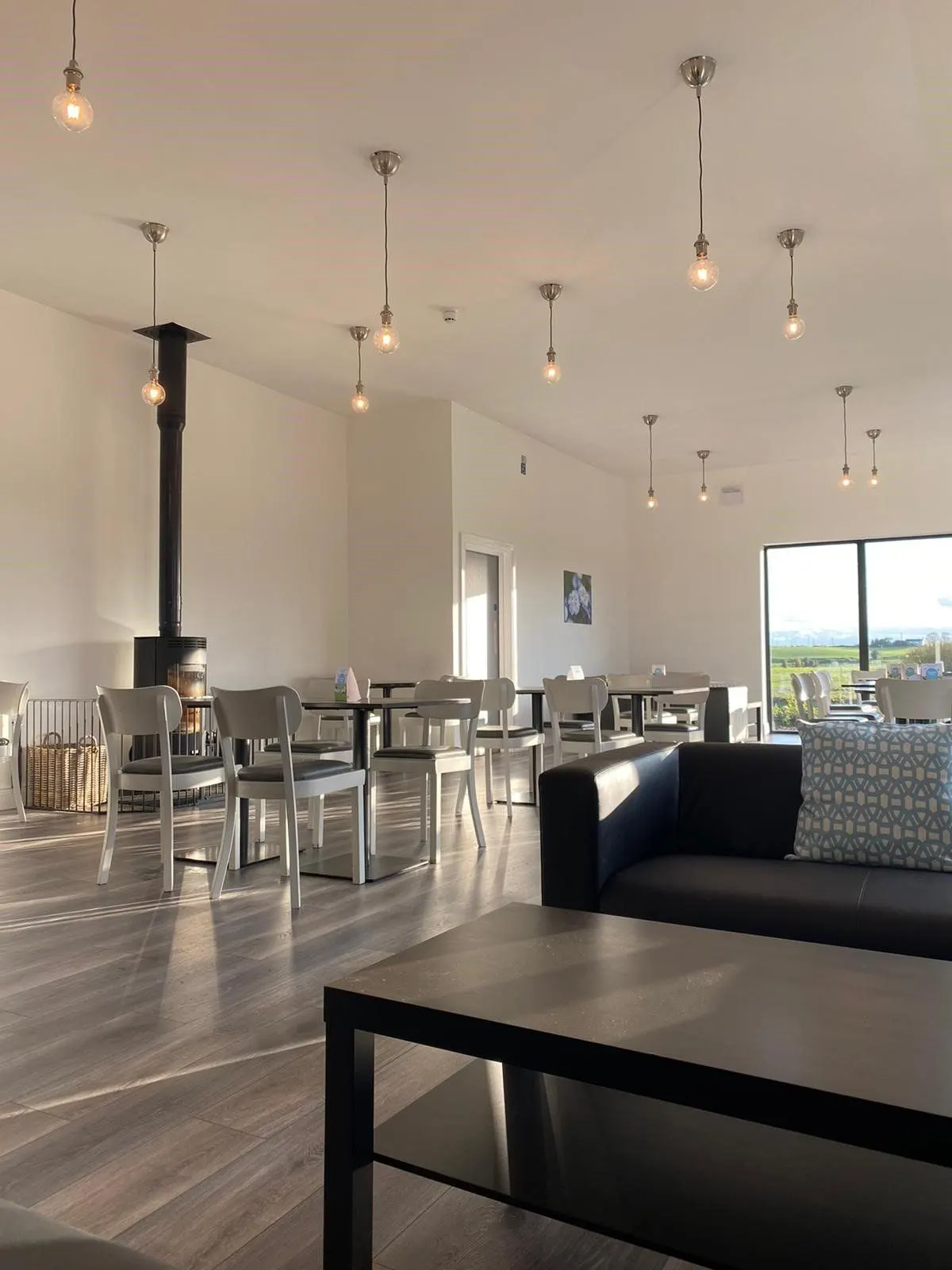 Modern open-plan leisure venue with pendant lights, seating areas, wood flooring, a black wood-burner, and countryside views through large windows.