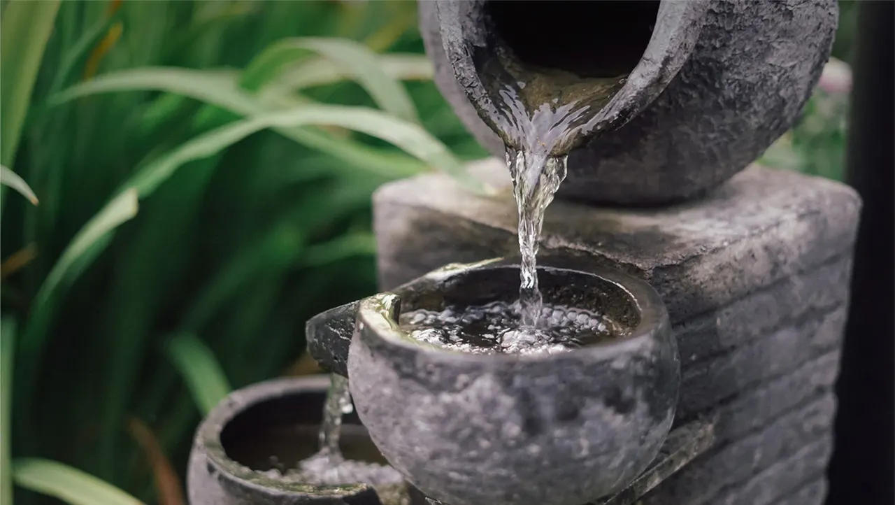 Water cascades from a stone spout into a tiered basin surrounded by green foliage.