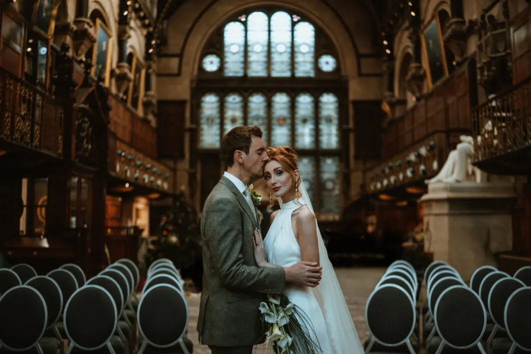 Bride and groom posing together in a grand historic hall with ornate arched windows and stone columns.