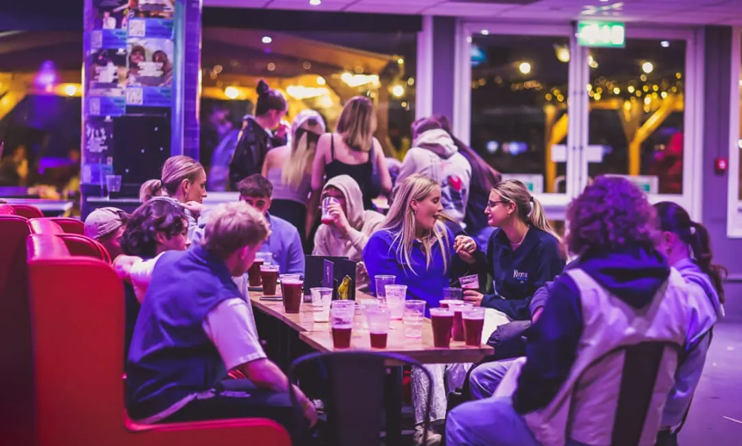 Groups of people socialising at tables with drinks in a vibrant purple-lit indoor leisure venue.