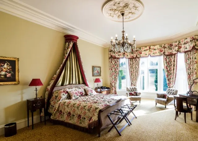 Elegant bedroom with four-poster bed, burgundy canopy, floral bedding and curtains, chandelier, and garden views through large windows.