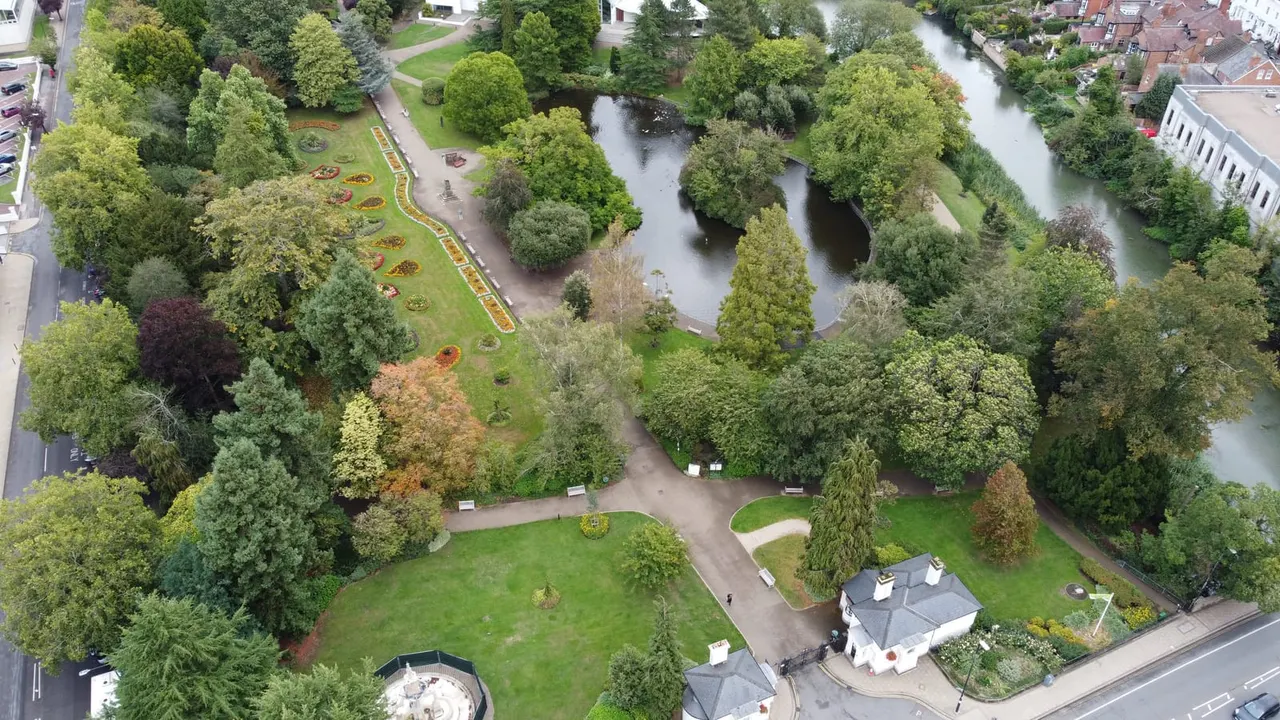 Aerial view of formal gardens with mature trees, flower beds, riverside paths and surrounding residential buildings.