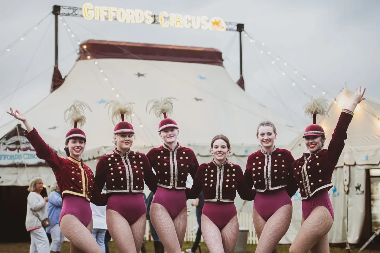 Six circus performers in matching burgundy and gold costumes with feathered hats pose in front of a striped big top tent.