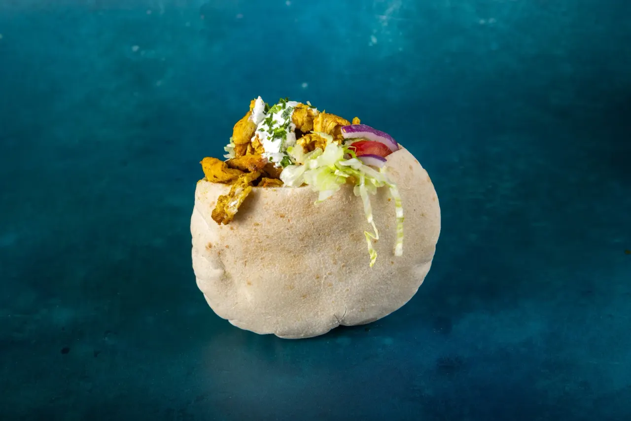 Fluffy pita bread filled with spiced cauliflower, slaw, tomato and sour cream on a teal background.