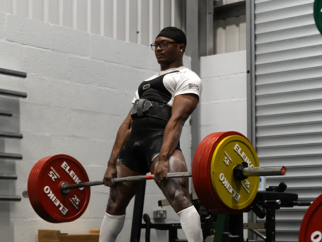 A man in athletic wear and protective gear performs a barbell lift with red and yellow weighted plates in an indoor gym.