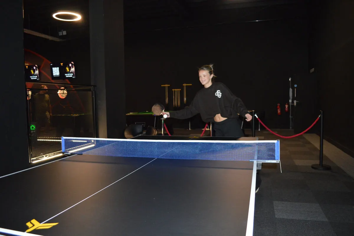 Woman playing table tennis indoors at an entertainment venue with arcade games visible in background.