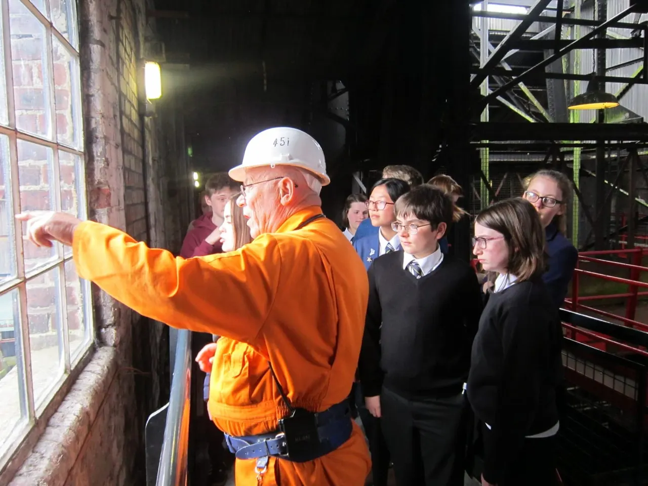 A tour guide in orange safety clothing and hard hat points at displays while talking to a group of young people during a night-time museum visit.