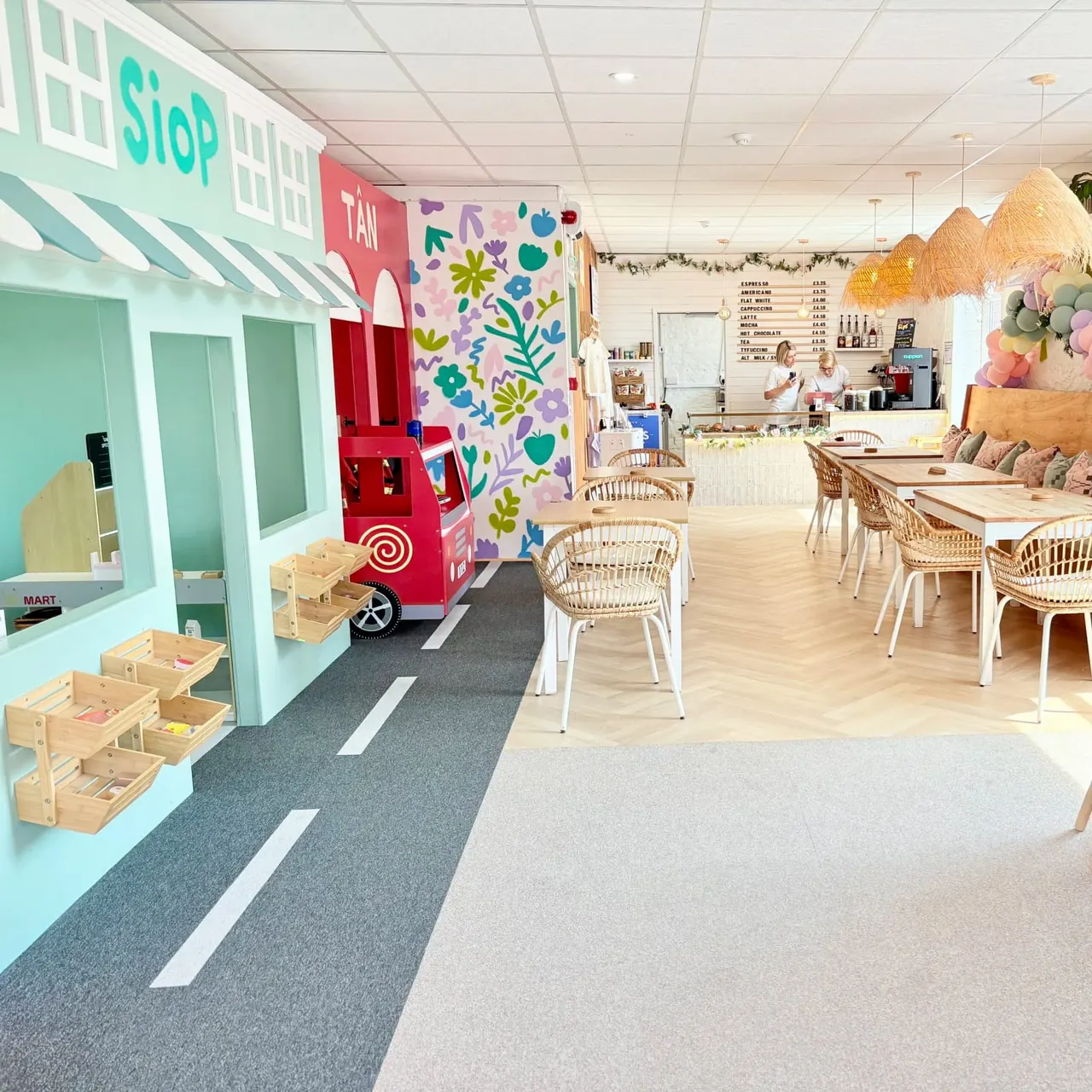 Colourful indoor play venue with toy shop and food truck props, wicker chairs, tables, and staff member behind counter.