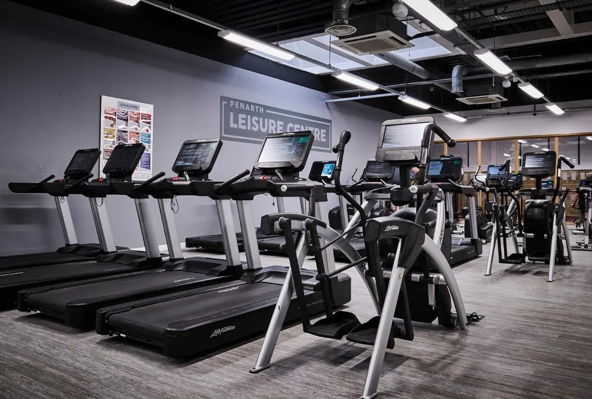 Modern gym interior with rows of treadmills and exercise bikes with digital screens, under bright ceiling lights.