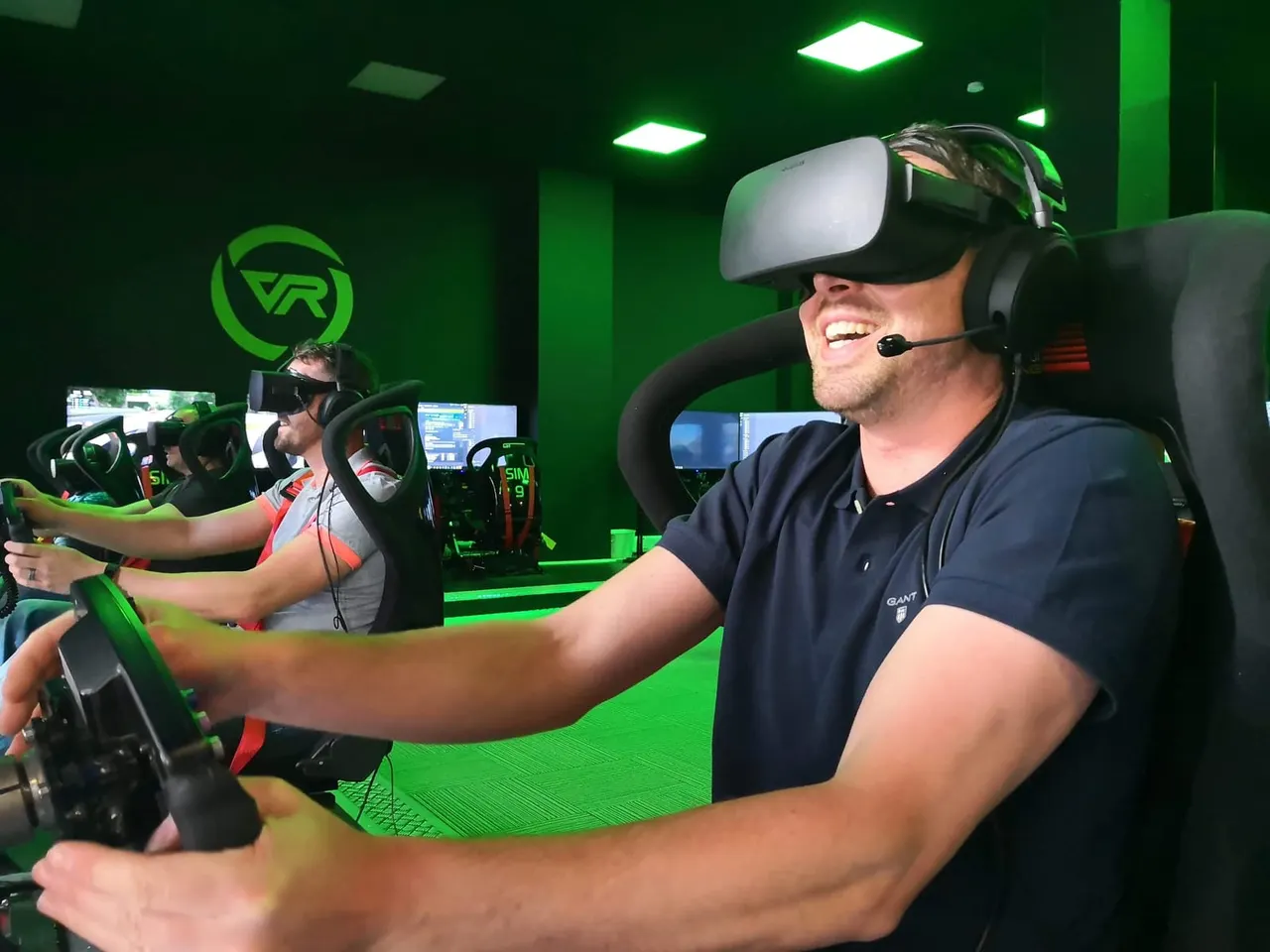Two people wearing VR headsets operate racing simulators in a green-lit arcade space.