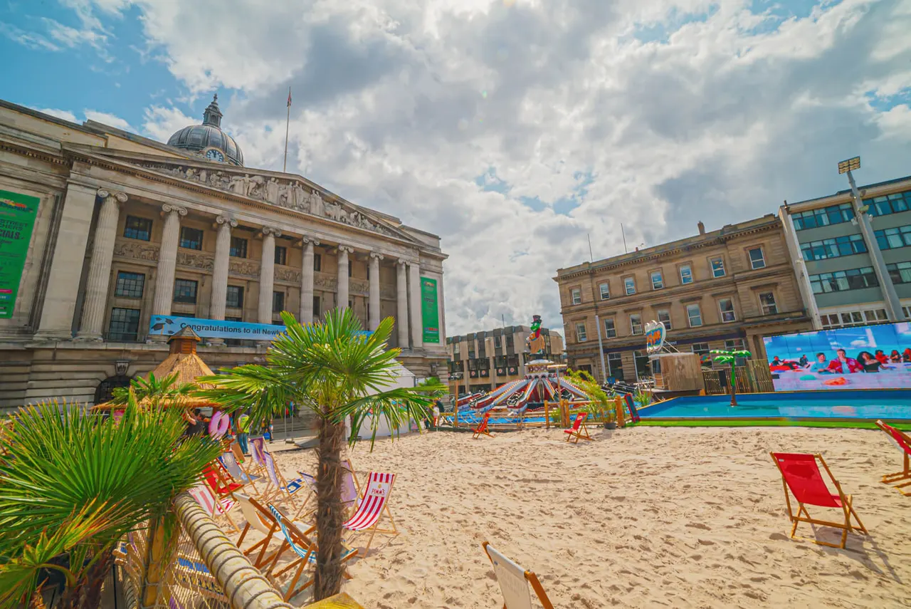 Temporary beach venue in city square with sandy area, deckchairs, palm trees and inflatable water slide beside classical buildings.