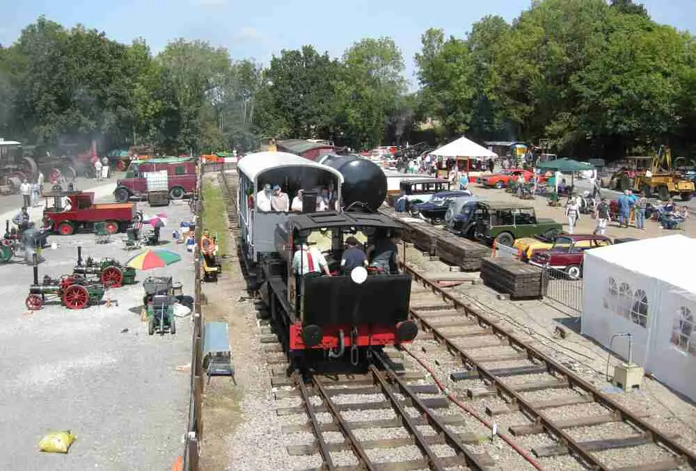 A heritage steam locomotive pulling a passenger carriage along narrow-gauge railway tracks, with vintage farm machinery and visitors visible at the station around it.
