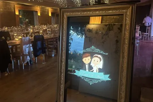 Elegant event space with framed digital display showing illustrated couple, guests dining at tables in background.