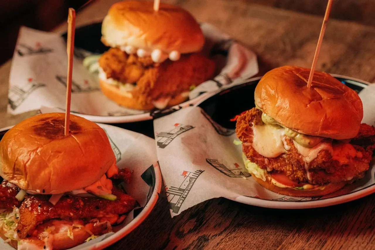 Three fried chicken sandwiches with glossy brioche buns, cheese and slaw on branded plates with cocktail picks.