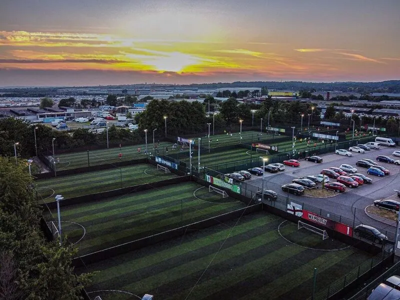 Aerial view of floodlit football pitches at sunset with parked cars and a waterside landscape beyond.