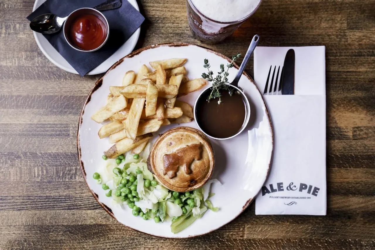 A plate of steak and ale pie with chips and peas, served with gravy, alongside cutlery and a soft drink.
