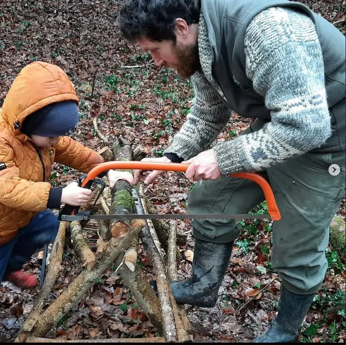 Adult and child using a saw to cut a branch together on a woodland floor covered with leaves.