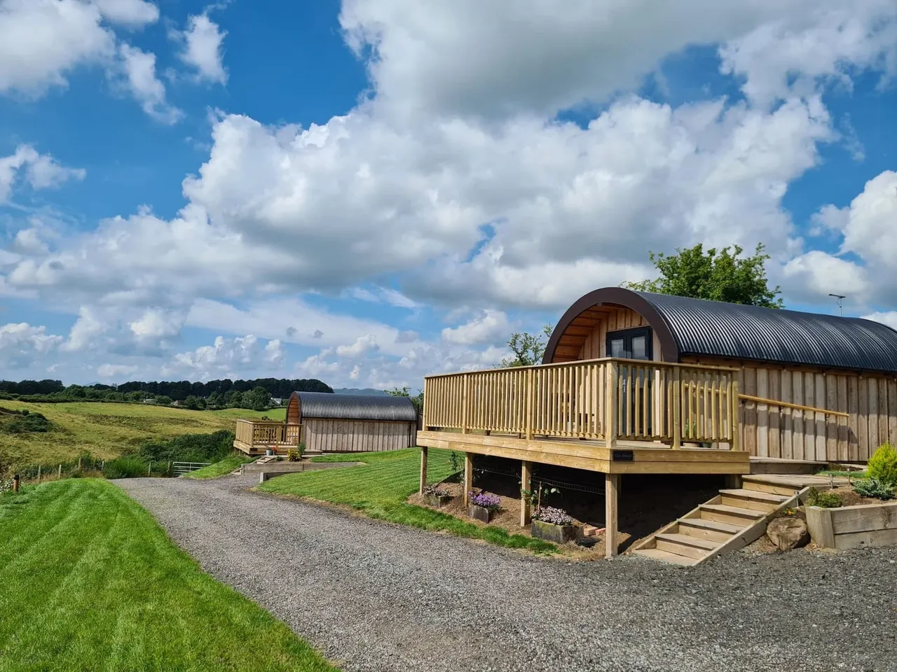 Glamping pods with curved metal roofs and wooden decking set in rolling countryside under a blue cloudy sky.
