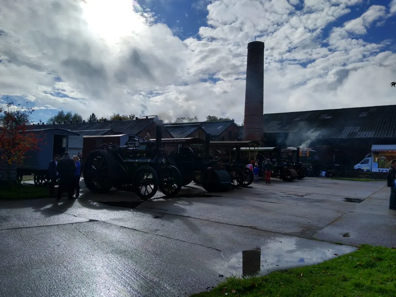 Visitors explore vintage steam engines and industrial machinery at an open-air heritage site with period buildings and a tall chimney.