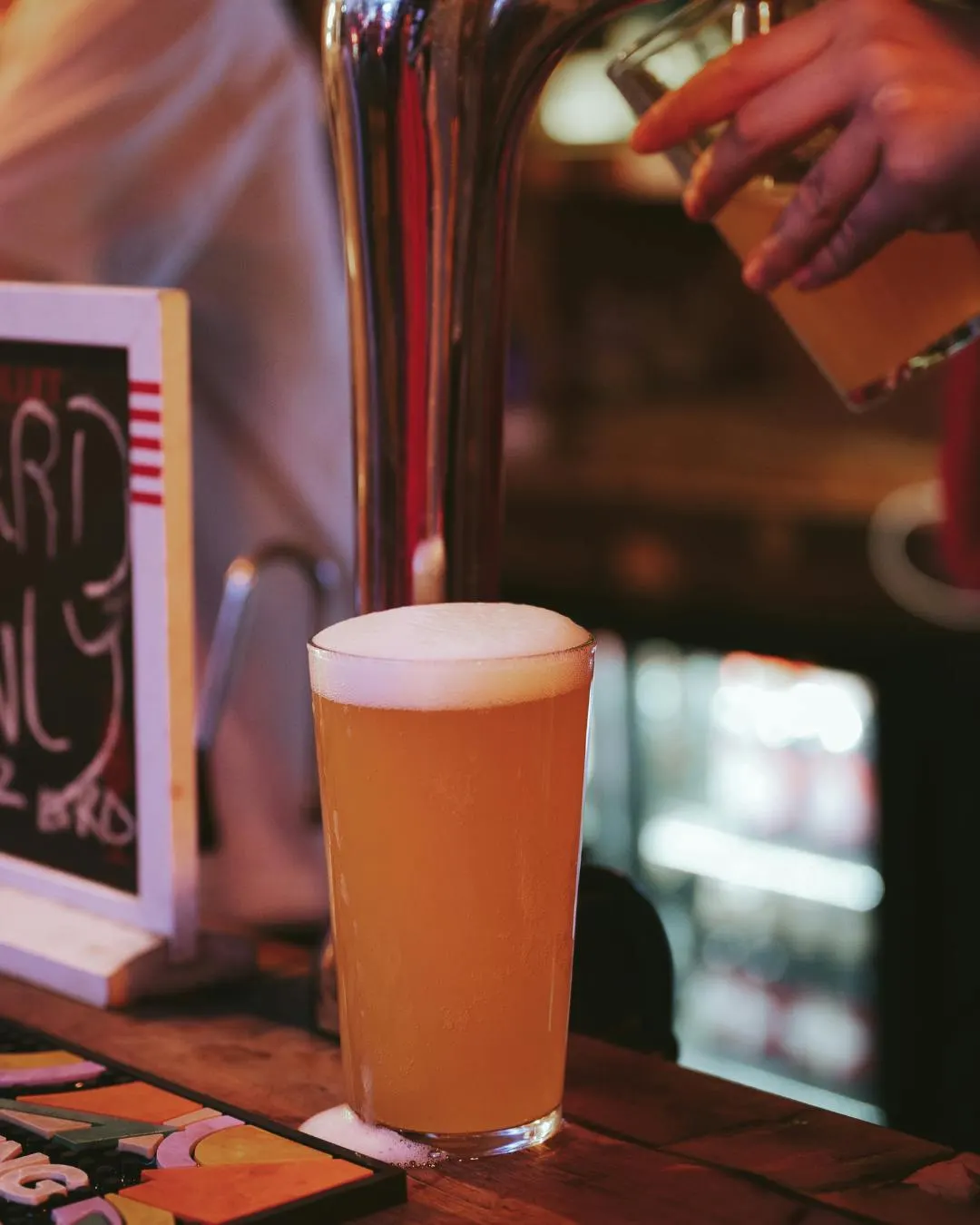 Pint of amber ale with creamy head on a bar counter next to beer taps and signage.