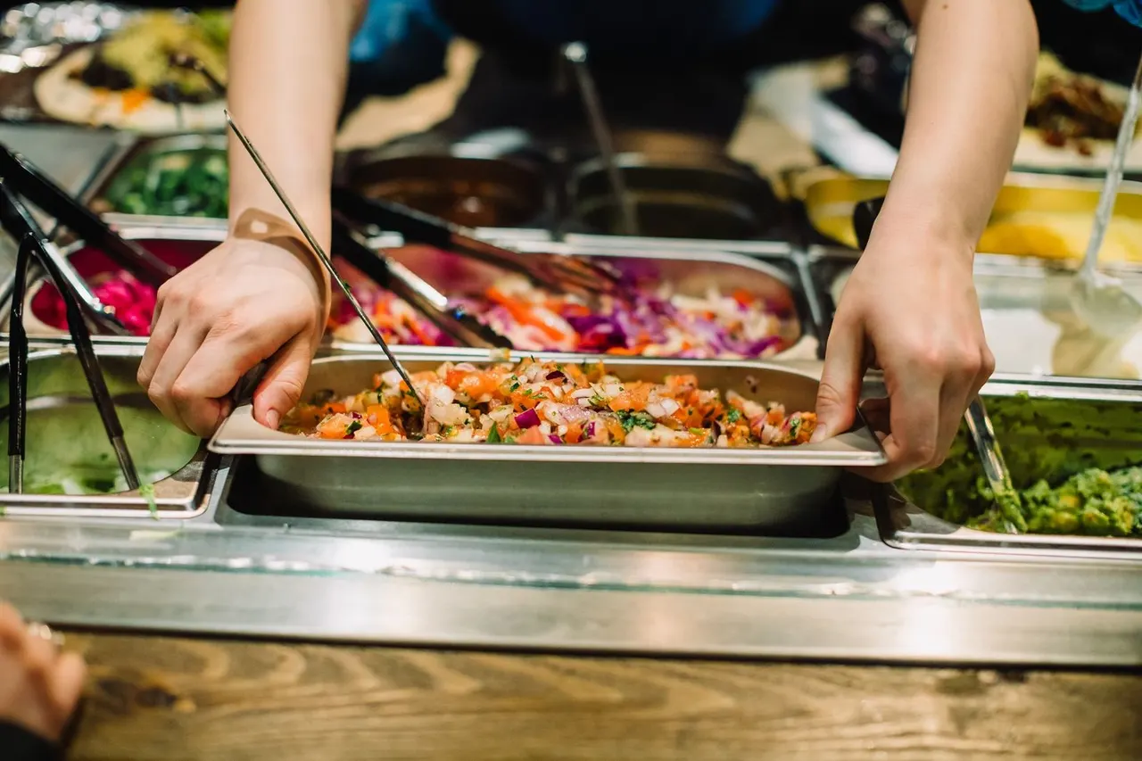 Person serving colourful mixed vegetable dish from a heated buffet counter with tongs.