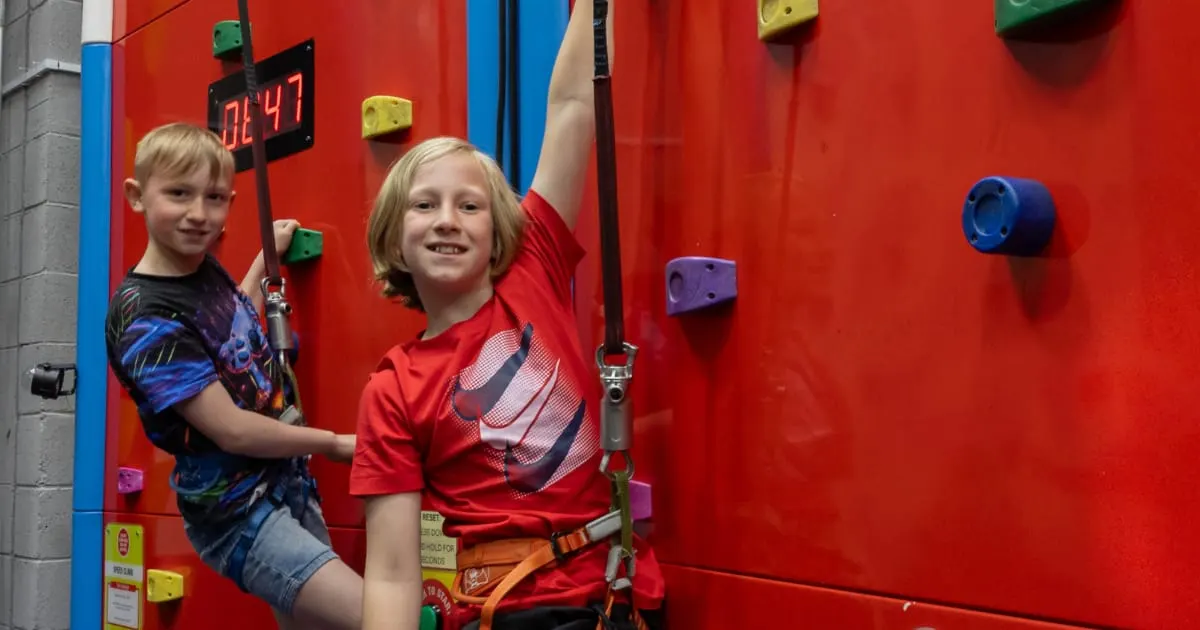 Two children climbing an indoor rock wall with coloured holds and safety harnesses.