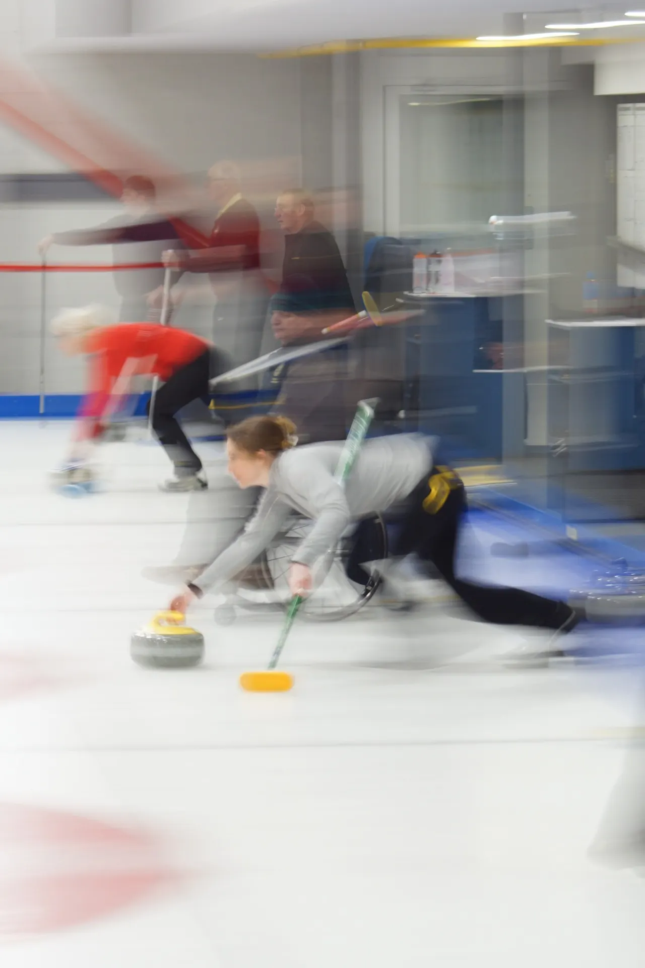 Curler in grey sliding across ice whilst releasing a stone during a match, with instructors and other players in background.
